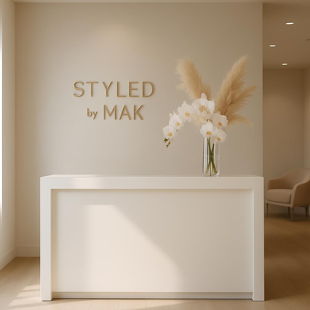 A serene, impeccably styled salon reception area featuring a minimalist white lacquer desk with clean lines, positioned in front of a pale greige wall bearing the subtle, metallic logo "Styled by Mak" in slender modern lettering. Behind the desk, a single tall glass vase holds an arrangement of white orchids and soft pampas plumes, adding delicate texture. Natural afternoon light streams in from an unseen window, blending with discreet recessed ceiling lights to create a soft, even glow and gentle shadows along the floor. Shot from a wide, eye-level perspective with balanced composition, the scene includes a hint of plush, light-taupe seating in the blurred background. The atmosphere is calm, exclusive, and welcoming, with photographic realism and a sophisticated, contemporary beauty studio aesthetic.